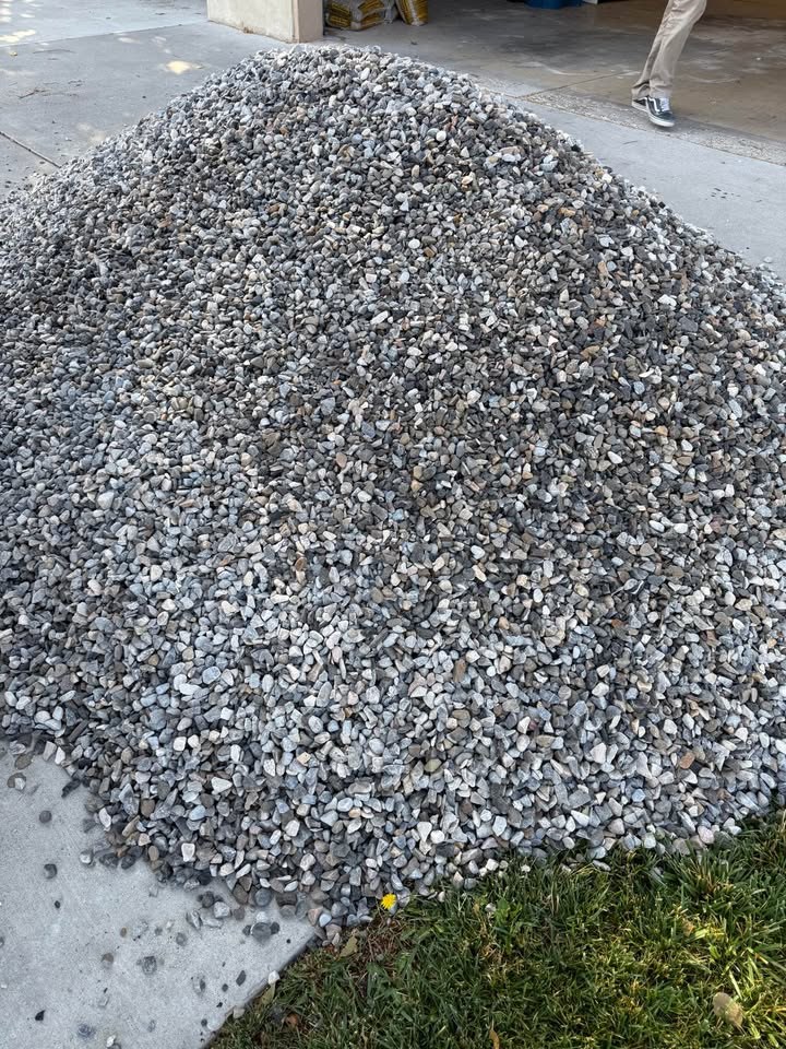 A large pile of grey drain rock dropped on a driveway apron, spilling just onto the grass