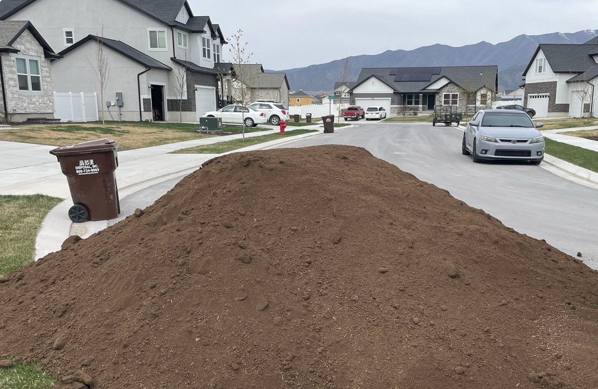 A long pile of screened topsoil dropped along a residential street, houses and mountains behind